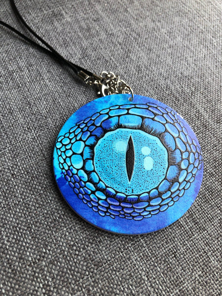 Collier breloque garçon rond bois modèle œil crocodile ou dragon fond bleu Comme un ange